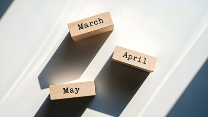 Wooden calendar blocks displaying the sequential months of march april and may resting on a bright white surface under dramatic light shadows