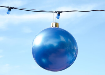 Giant Blue Christmas ball ornament hanging from black power line with small blue lights. Blue cloudy sky background.