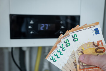 Hand holding fan of fifty euro banknotes in front of white gas boiler thermostat symbolizing high energy heating costs