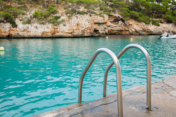 Metal pool ladder leading to turquoise sea water in Cala Santanyi bay on Mallorca, Spain. Close up of crystal clear waving water and cliffs over the sea