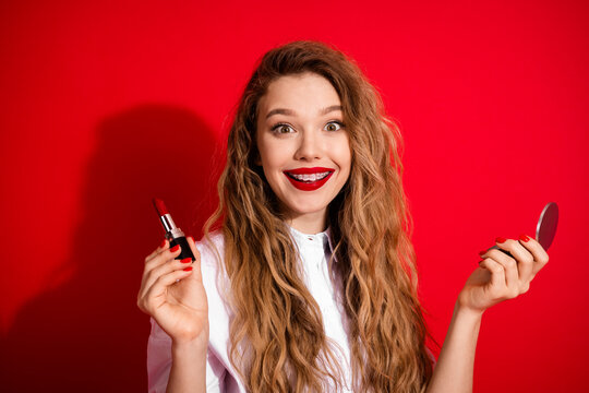 Cheerful woman holding red lipstick and mirror on a vibrant red background, showcasing beauty and style - Powered by Adobe