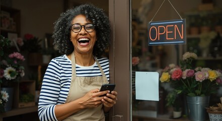 Happy mature african american woman florist laughing at her shop entrance. Successful small business owner with open sign holding smartphone