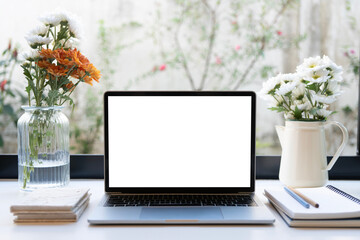 Laptop and notebook with flowers vase on white desk by the window