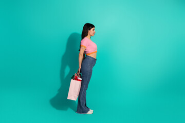 Young stylish brunette woman stands in profile with pink shopping bag against teal background