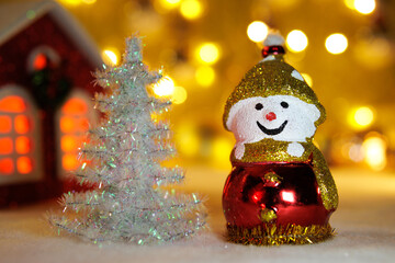 Against a blurred background with bright lights and a red house, a snowman with a white Christmas tree