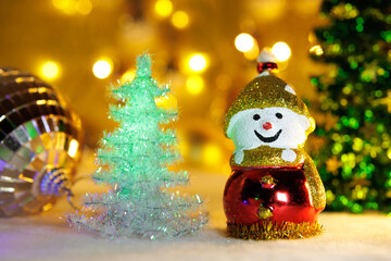 On a blurred background with bright lights are two Christmas trees with a large ball and a snowman