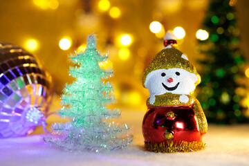 On a blurred background with bright yellow lights are two Christmas trees with a large ball and a snowman
