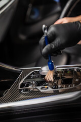 A gloved hand uses a small detailing brush to clean dust from a glossy center console with carbon fiber trim, a knurled metal dial, and a crystal like gear selector. © Aerial Film Studio