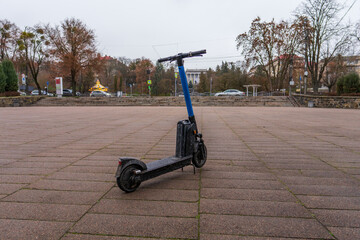 An electric scooter stands on the ground in an urban square. Trees and buildings are visible in the background on a cloudy day during the fall season © Роман Булатов