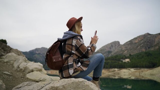 Woman hiker in hat and spring outfit with backpack relaxes on large rock by peaceful lake, capturing breathtaking views of mountains on smartphone while on an adventurous getaway
