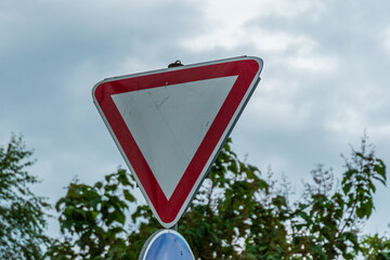 A triangular traffic sign with a red border and white center stands near a road surrounded by trees on a cloudy day. The sign signals drivers to yield