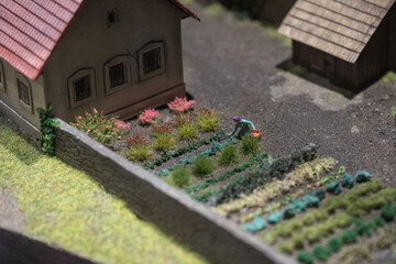 A mini figurine of a woman with a hoe working in garden beds in a model scene.
