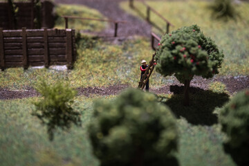 A mini figurine of a man picking apples from a tree with a ladder in a model scene.
