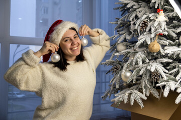 Woman in Santa hat holding ornaments by a Christmas tree indoors.