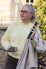 Confident woman in yellow sweater enjoying a sunny day while holding a phone and a jacket in a charming outdoor setting