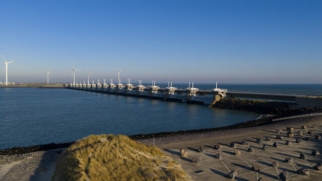 Aerial view of the formidable Oosterscheldekering barrier stretching across the water, contrasting with the soft dunes and clear sky, Vrouwenpolder, Zeeland, Netherlands.