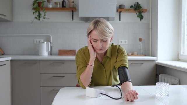 Exhausted middle aged woman with signs of headache and low blood pressure measuring pressure with tonometer while sitting in kitchen. Female feeling unwell, having migraine and signs of sickness.