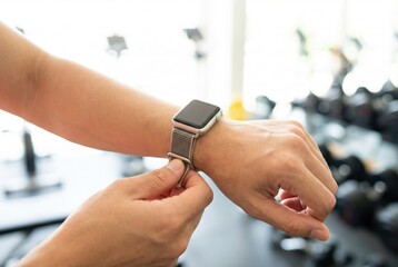 Person adjusts fitness tracker on wrist in gym during workout session with equipment in background and natural light coming through windows