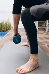 Athletic individual performing a strength training exercise with a blue dumbbell on a yoga mat beside a swimming pool, showcasing fitness and dedication to health