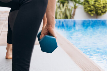 Athletic woman in black leggings is exercising with a blue dumbbell beside a swimming pool, showcasing strength and determination in a vibrant outdoor setting