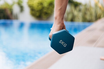 Individual holding a blue hexagonal dumbbell labeled 5KG, positioned near a serene swimming pool, showcasing fitness and wellness lifestyle in a vibrant outdoor setting