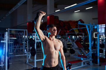 Muscular athlete performing one-arm overhead kettlebell press in a gym.