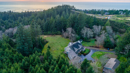 Aerial view of a cozy house nestled amidst towering evergreens, with the distant ocean horizon painting a soft glow, Tillamook, Oregon, United States.