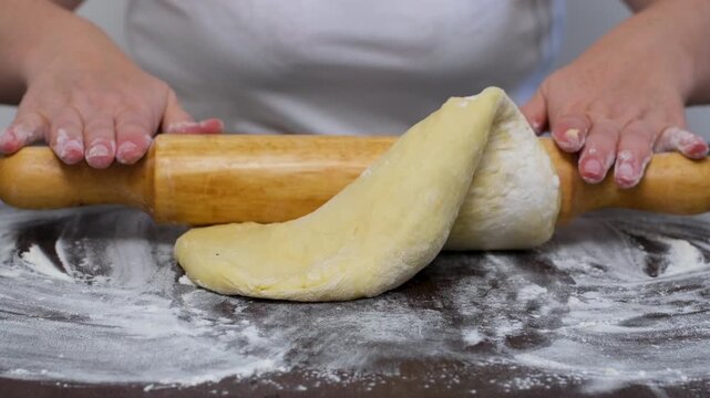 Woman baker is rolling out dough using wooden rolling pin on cozy wooden table. She is rolling dough in flour, preparing it for an Italian pizza with tomato and cheese.