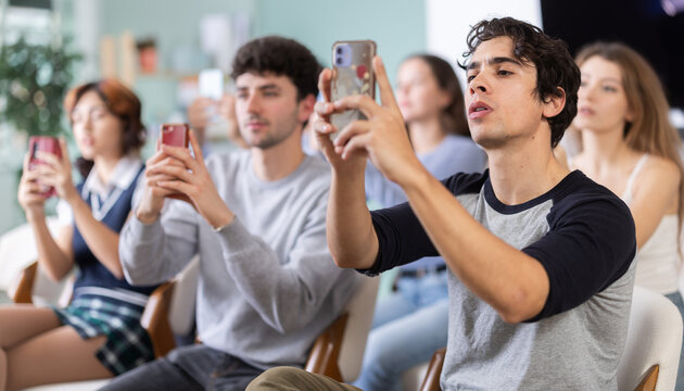 Young man holds a mobile phone and takes photos of study materials during a lecture in class. Students scan the barcode to view the entire lecture on the website - Powered by Adobe