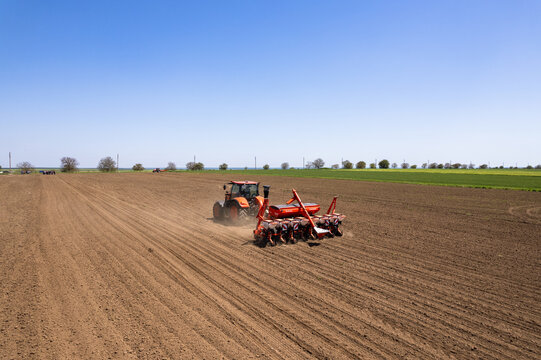 Aerial view of a vibrant orange tractor contrasts against the rich brown earth, preparing the land for planting under a vast blue sky, Silistra, Bulgaria.