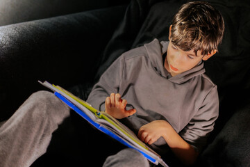 Young boy with short hair, wearing a gray hoodie, is reading a colorful book while sitting comfortably on a dark couch, immersed in the world of stories and imagination