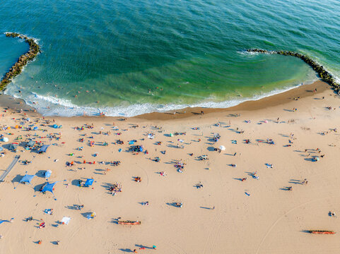 Aerial view of sunbathers dotting the sandy expanse where the beach meets the foamy, turquoise waves guarded by rocky jetties, Brooklyn, New York, United States.