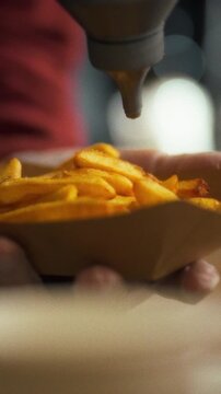 Skilled cook in busy kitchen gently pours rich culinary sauce over gourmet fries served in simple paper holder, showcasing food's vibrant colors and diverse textures, elevating the dining experience