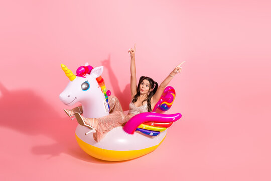 Young woman in glitter dress enjoys a playful ride on a colorful unicorn inflatable float against pink background with a fun fashion party vibe