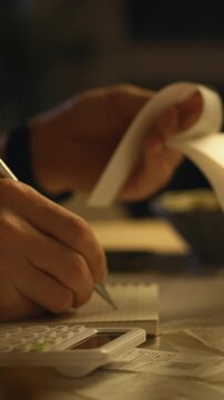 In dimly lit workspace, individual diligently sorts receipts uses calculator to manage finances at night, creating detailed budget through meticulous accounting expense tracking their cluttered desk
