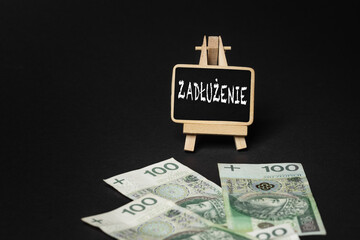 Black wooden writing board on a black background with the inscription "Zadłużenie", in the background scattered Polish złoty PLN banknotes (selective focus)