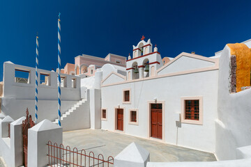 White church with red bell tower and Greek flags in Oia village on Santorini island, Greece, under a clear blue sky.