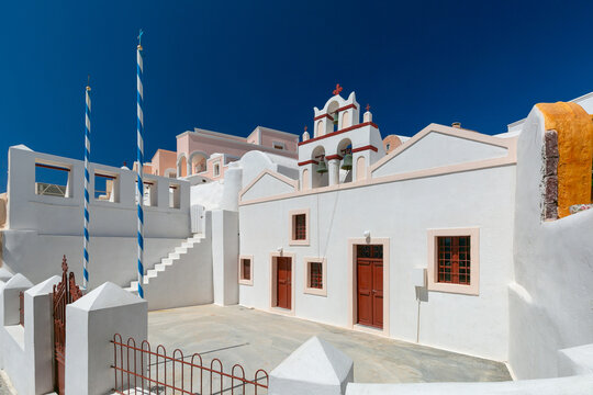 White church with red bell tower and Greek flags in Oia village on Santorini island, Greece, under a clear blue sky. - Powered by Adobe