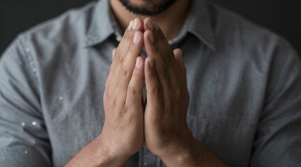 Person clasps hands in reverence, wearing a grey button down shirt against dark background