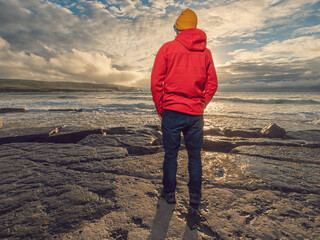 A man in a red jacket stands on a rocky beach, with the sun setting in the background. Scene is peaceful and serene, as the man is enjoying the beauty of the natural surroundings. Doolin, Ireland