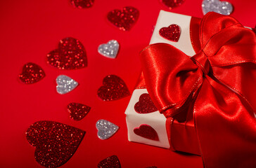 White Gift Box wrapped with a luxurious red satin bow, decorated with glitter hearts on a Red. Festive holiday composition symbolizing love, celebration, surprise, and special occasions, with cop