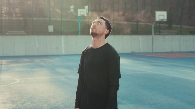 A serious young man in a black tee stands on a sun-drenched outdoor court. Intense gaze and focused expression under a warm direct sun light light. Pissed expression followed by a facepalm.