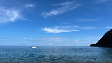 Small white boat floating on a calm blue sea with wispy clouds, mountains, and a tropical coastline