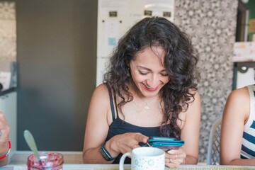 A young Latina woman eating a healthy breakfast, looking at her cell phone and sending a text message, sitting in the dining room of her modern apartment. Healthy lifestyle and nutrition concept.