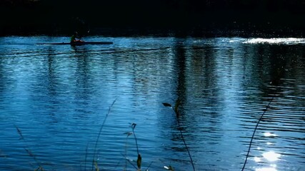 Silhouette of a man kayaking on a calm blue lake. Silhouetted man paddling a kayak on a serene blue lake, with sunlight glittering on the water's surface