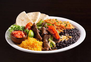 Tex-Mex steak fajita platter with seared beef strips, rice, black beans, salad and flour tortillas in a white plate on a dark surface; copy space