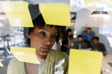 Focused black young businesswoman writing ideas on yellow sticky notes on a glass board while actively brainstorming and planning a new business strategy
