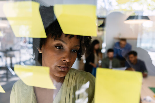 Focused black young businesswoman writing ideas on yellow sticky notes on a glass board while actively brainstorming and planning a new business strategy - Powered by Adobe
