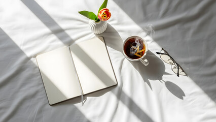 An open notebook, a cup of steaming tea, a single tulip, and glasses are neatly arranged on white bed linen bathed in morning sunlight.