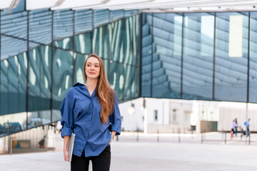 Confident young businesswoman in blue shirt with laptop standing near modern glass building with stylish and professional appearance during daytime